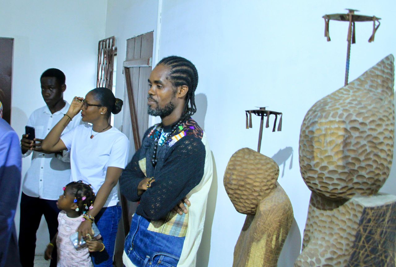 Plusieurs personnes observent des sculptures en bois exposées dans une galerie aux murs blancs