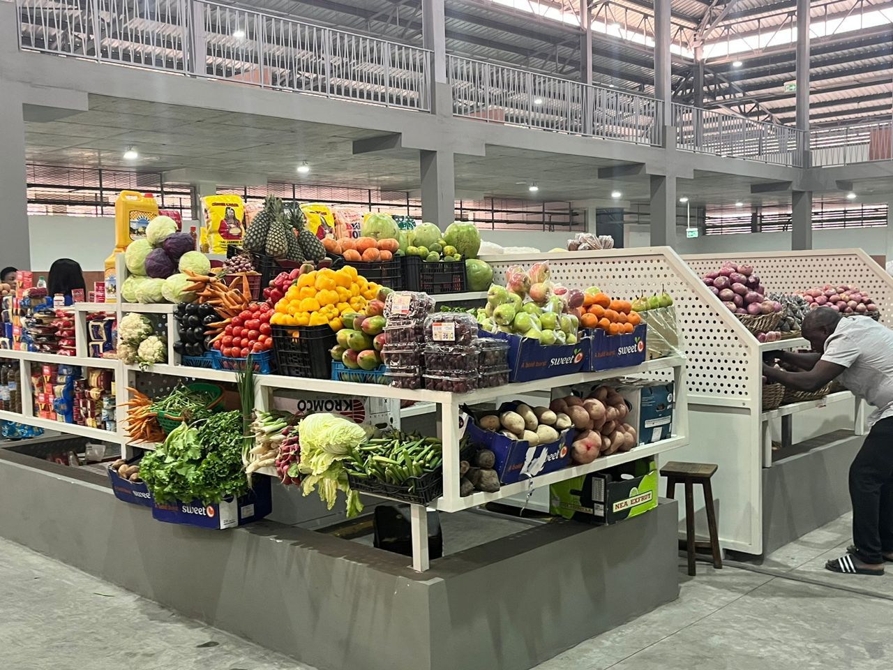 Étal de marché couvert avec fruits et légumes frais colorés exposés et un homme rangent des oignons dans un casier