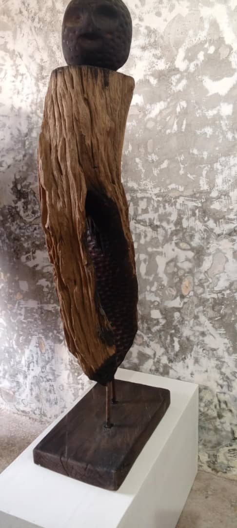 Sculpture en bois partiellement brûlé montée sur un socle blanc devant un mur texturé clair