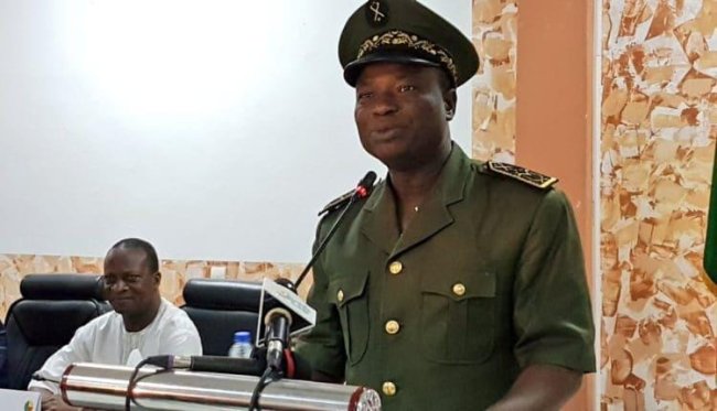 Un homme en uniforme militaire parle derrière un pupitre avec un microphone, un autre homme est assis en arrière-plan