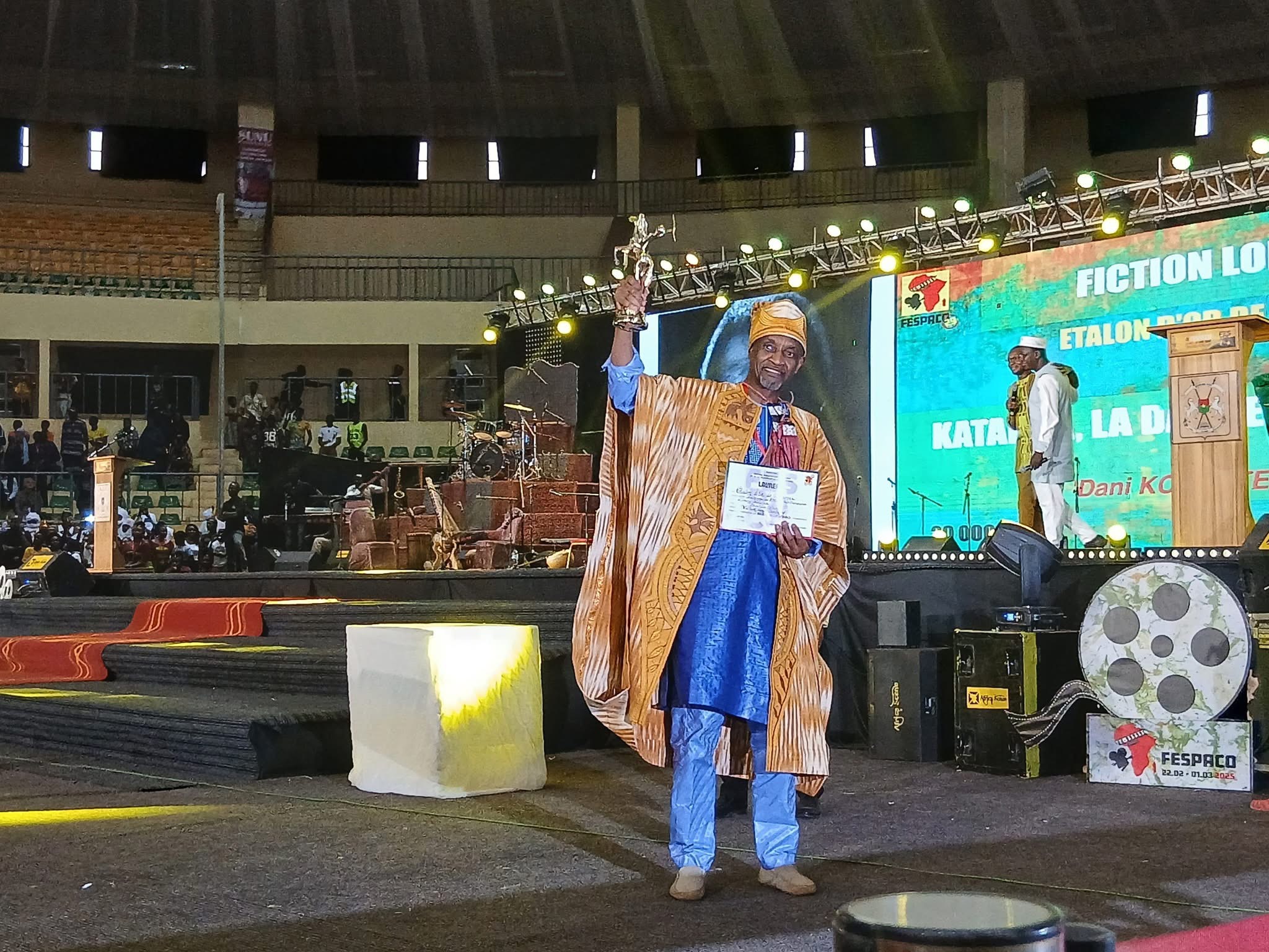 Un homme en tenue traditionnelle tient un trophée et un certificat sur une scène devant un écran lumineux affichant un titre de film