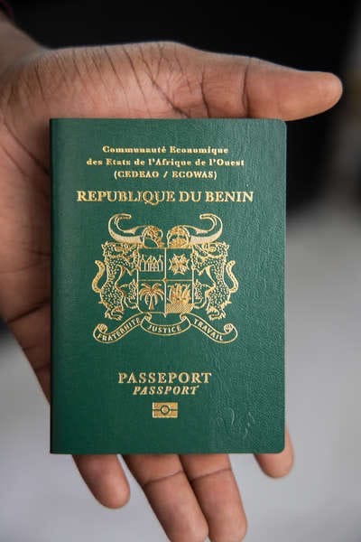 Une main tient un passeport vert foncé avec inscriptions dorées "République du Bénin" et un blason au centre