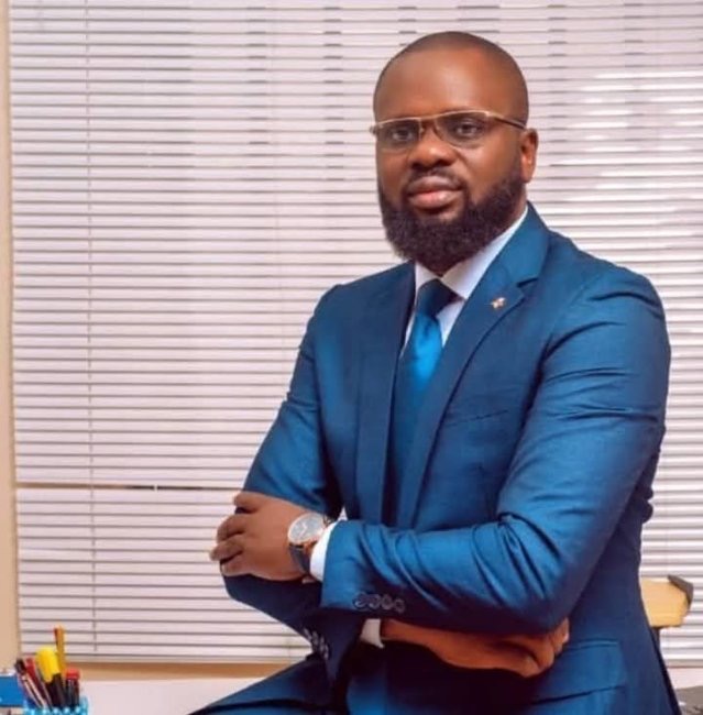 Un homme en costume bleu et lunettes pose assis devant un bureau devant des stores blancs fermés