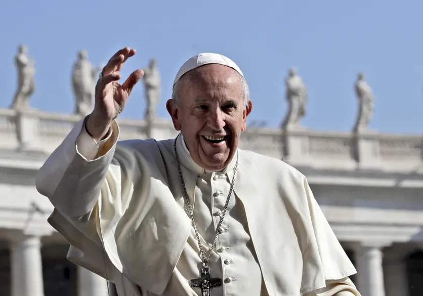 Un homme en tenue religieuse blanche agite la main devant un bâtiment décoré de statues sous un ciel bleu