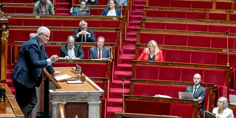 Un homme debout parle au micro devant un hémicycle aux sièges rouges avec plusieurs personnes assises l'écoutant attentivement
