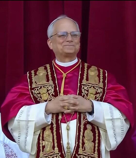 Homme vêtu d’une tenue religieuse richement décorée, sourire aux lèvres, devant un rideau rouge
