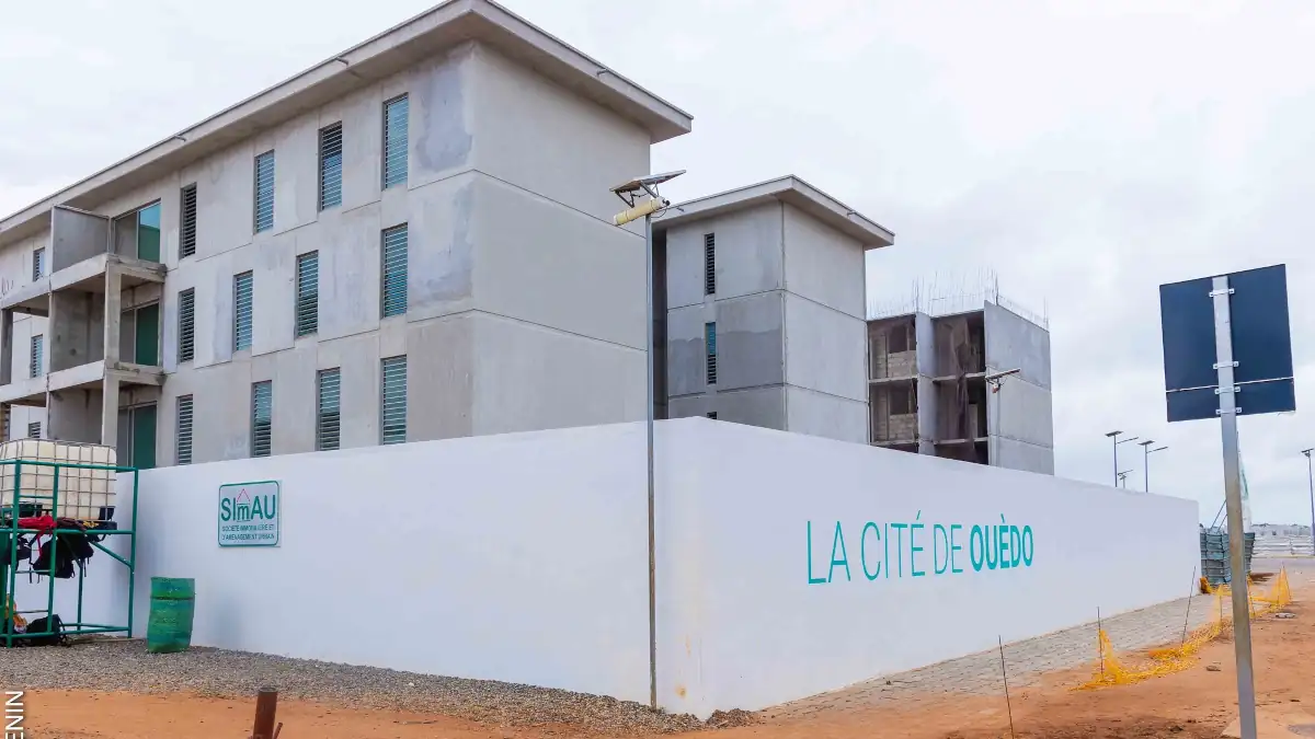 Bâtiments résidentiels inachevés derrière un mur peint indiquant La Cité de Ouédo sur un sol sablonneux
