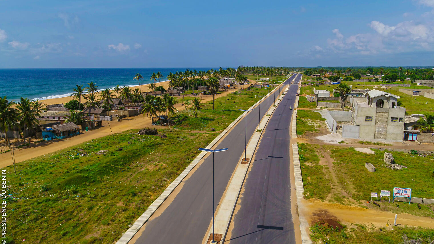 Route asphaltée bordée de pelouses et quelques bâtiments en construction près d une côte bordée de palmiers