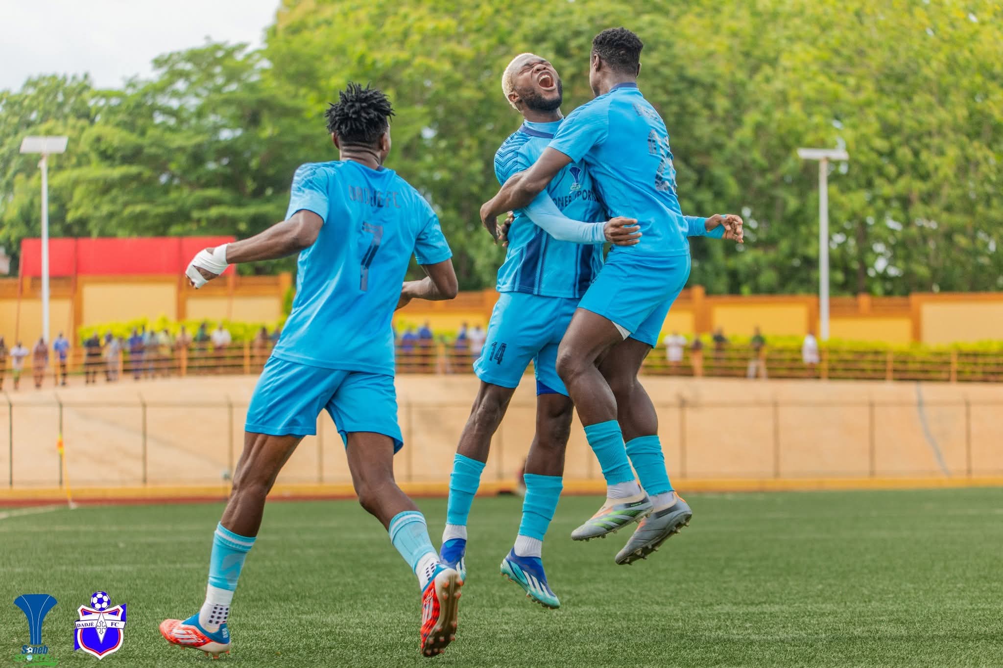 Trois joueurs de football en maillot bleu célèbrent ensemble un but sur un terrain en plein air avec des arbres en arrière-plan