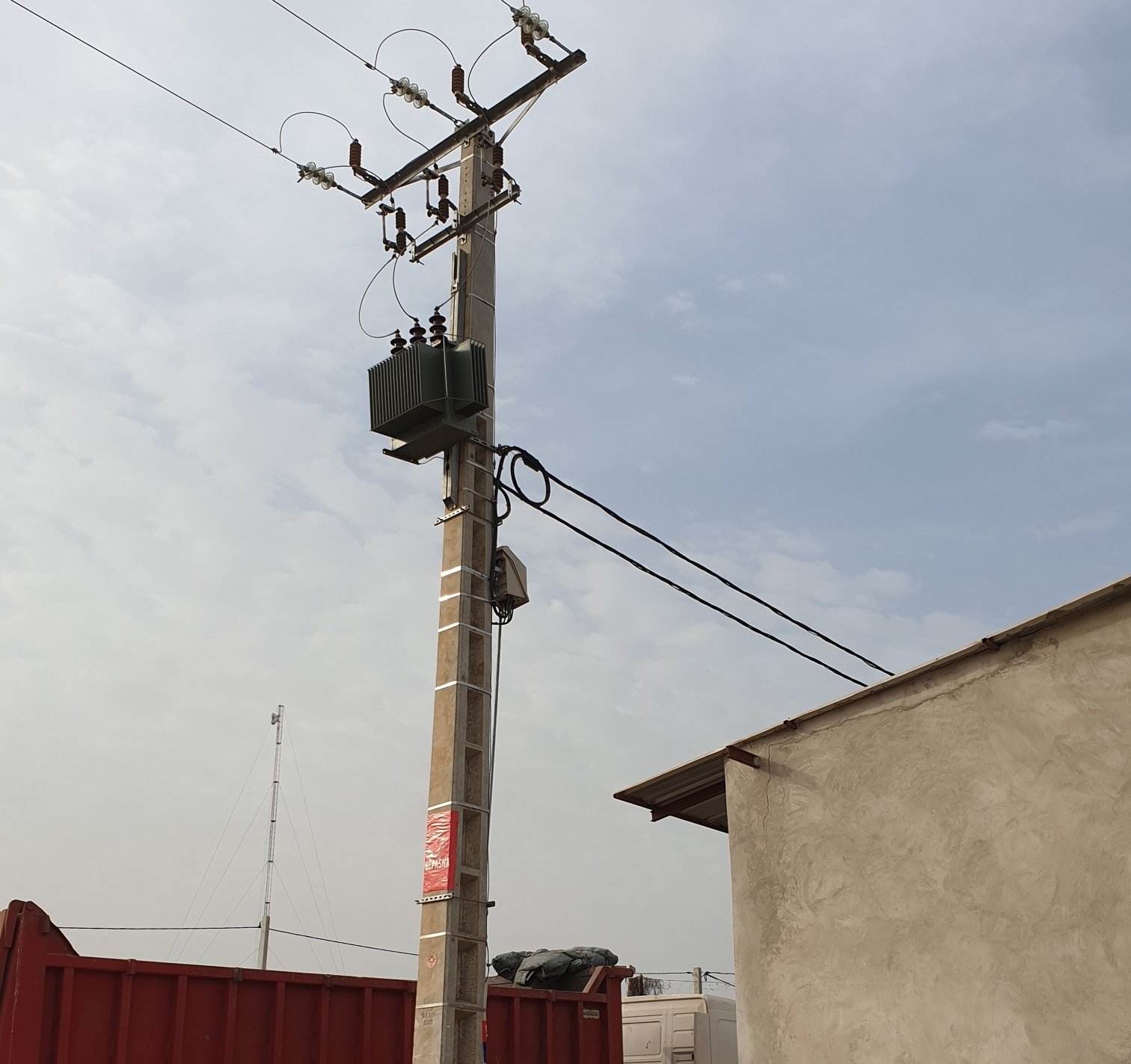 Poteau électrique avec transformateur surmonté de câbles sous un ciel gris clair près de bâtiments et containers rouges