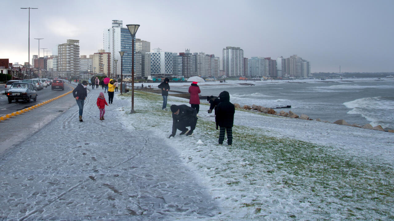 Personnes marchant et jouant sur un sol partiellement couvert de neige près d'une plage et de bâtiments en bord de mer