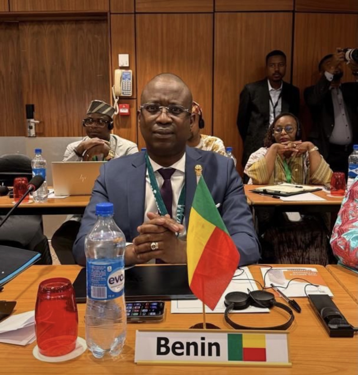 Homme en costume assis derrière une table avec un drapeau et une pancarte Benin entouré de plusieurs personnes dans une salle de réunion