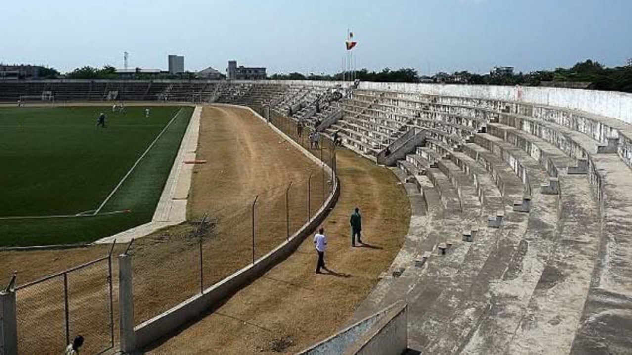 Stade avec tribunes en béton vieilles et pelouse entourée d'une piste, quelques personnes marchant autour du terrain