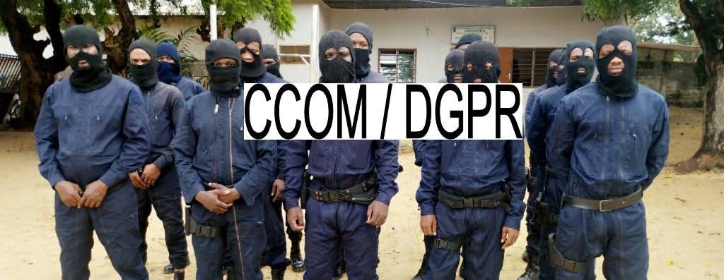 Homme en uniforme sombre avec cagoule noire formant un groupe debout en plein air devant un bâtiment clair