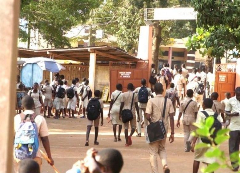Des élèves en uniforme beige marchent vers l’entrée d’un établissement scolaire sous des arbres ensoleillés