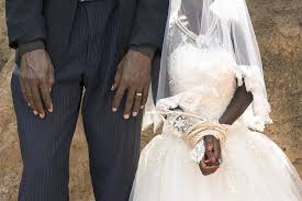 Un couple en habits de mariage se tient côte à côte devant un mur en terre battue, les mains visibles.