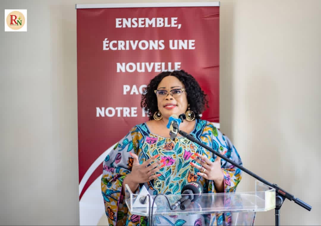 Une femme souriante en tenue colorée parle au micro derrière un pupitre transparent devant une banderole rouge texte blanc