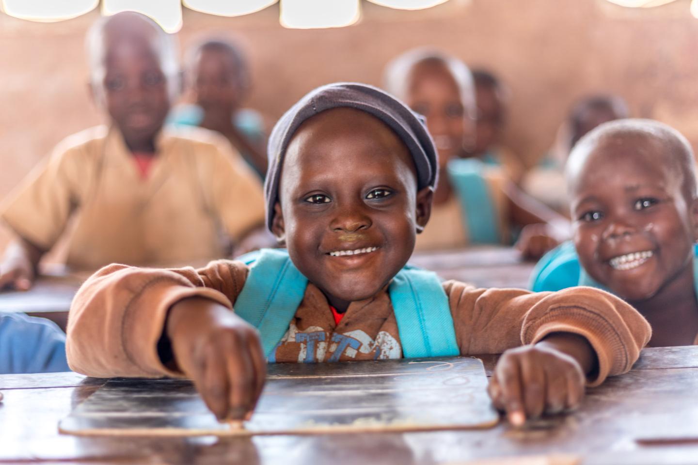 Plusieurs enfants souriants assis à des tables dans une classe éclairée par la lumière naturelle