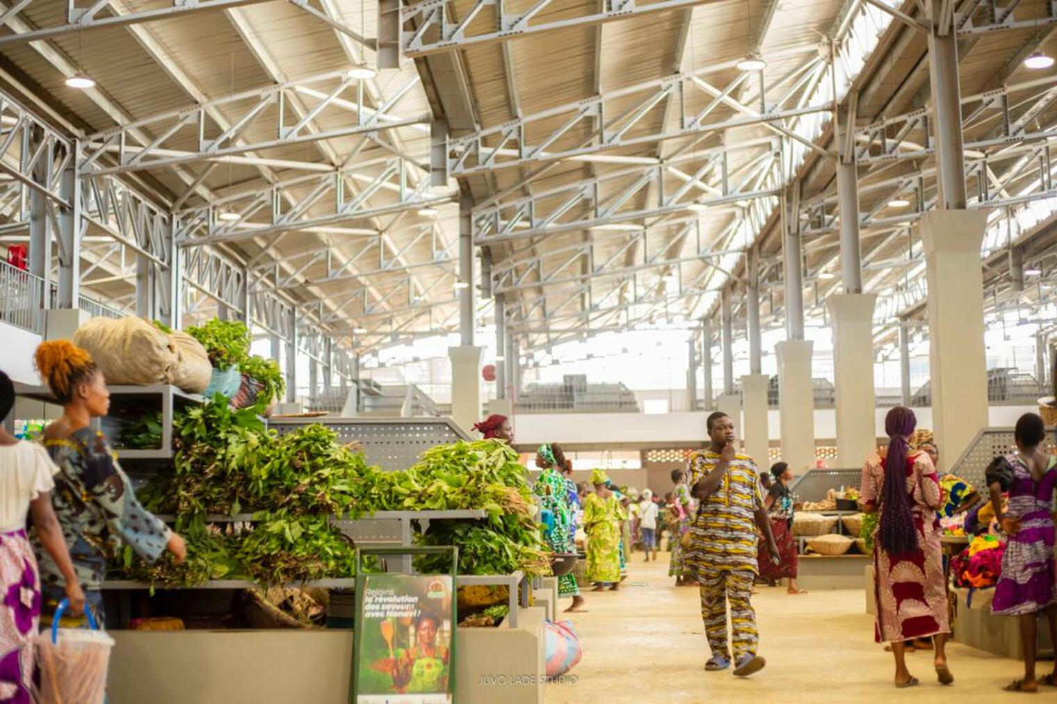 Marché couvert animé avec des étals de légumes et des personnes vêtues de pagnes colorés sous une grande toiture métallique lumineuse