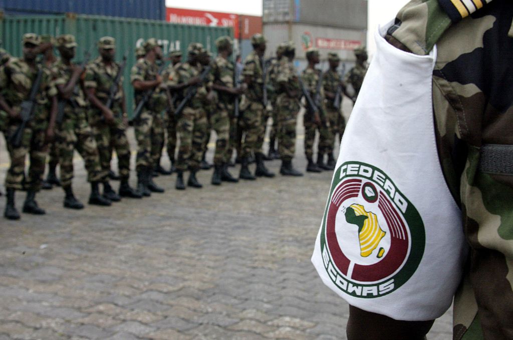 Soldats en uniforme de camouflage alignés en formation avec un brassard blanc portant un logo circulaire représentant l’Afrique et des inscriptions vertes et ro