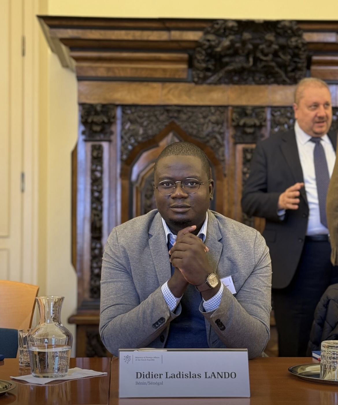 Un homme en veste grise assis à une table devant une plaque nominative lors d'une réunion dans une salle ornée.