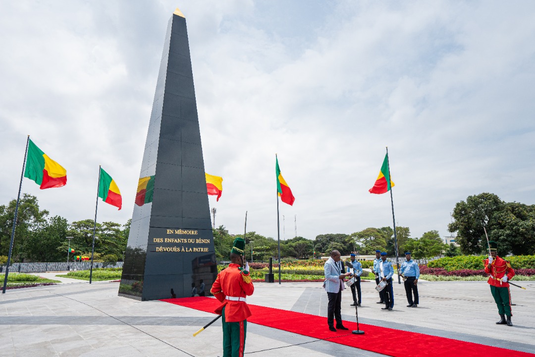 Un monument noir entouré de drapeaux tricolores rouges, jaunes, verts avec des militaires en uniforme et un homme au micro sur tapis rouge