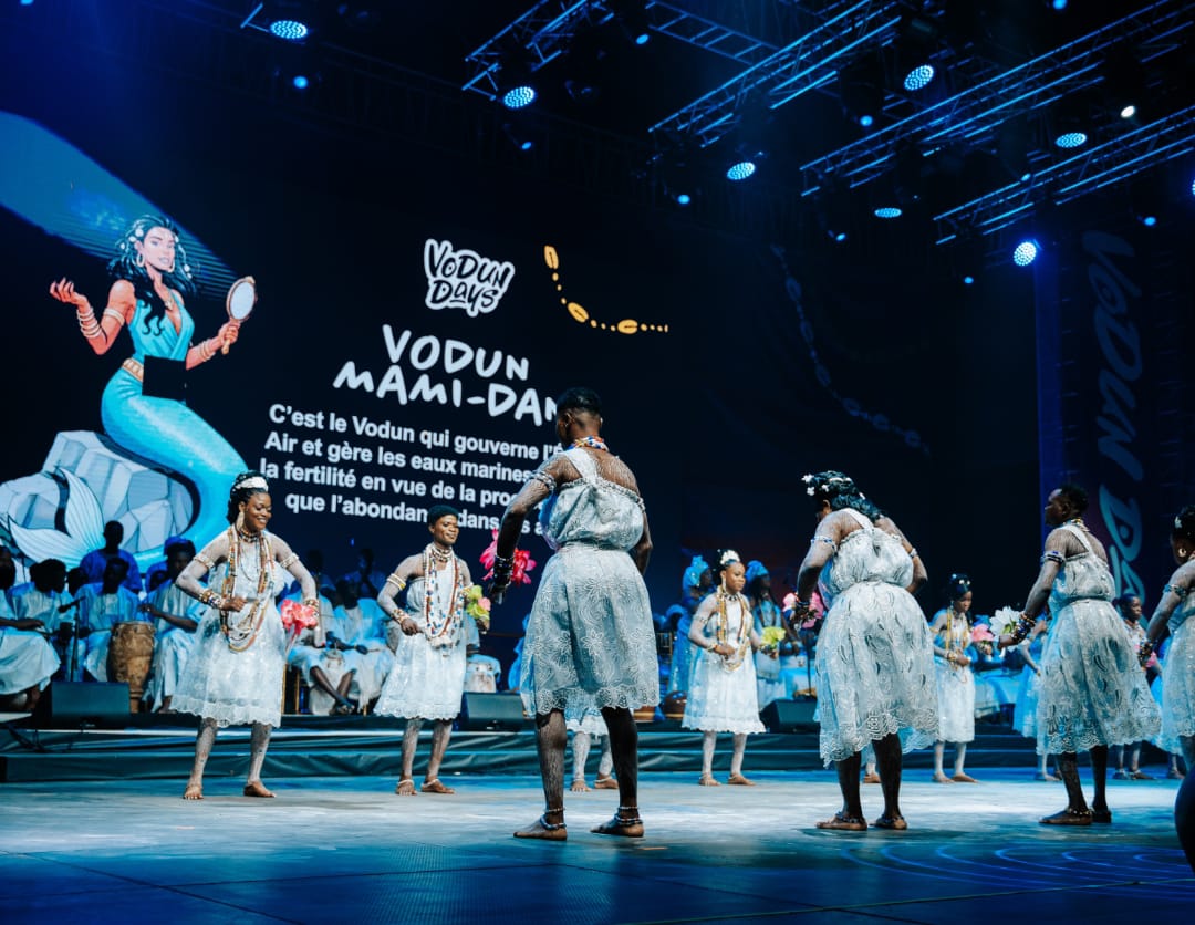 Des danseuses en robes blanches dansent sur une scène éclairée avec un écran montrant un dessin et du texte en arrière-plan