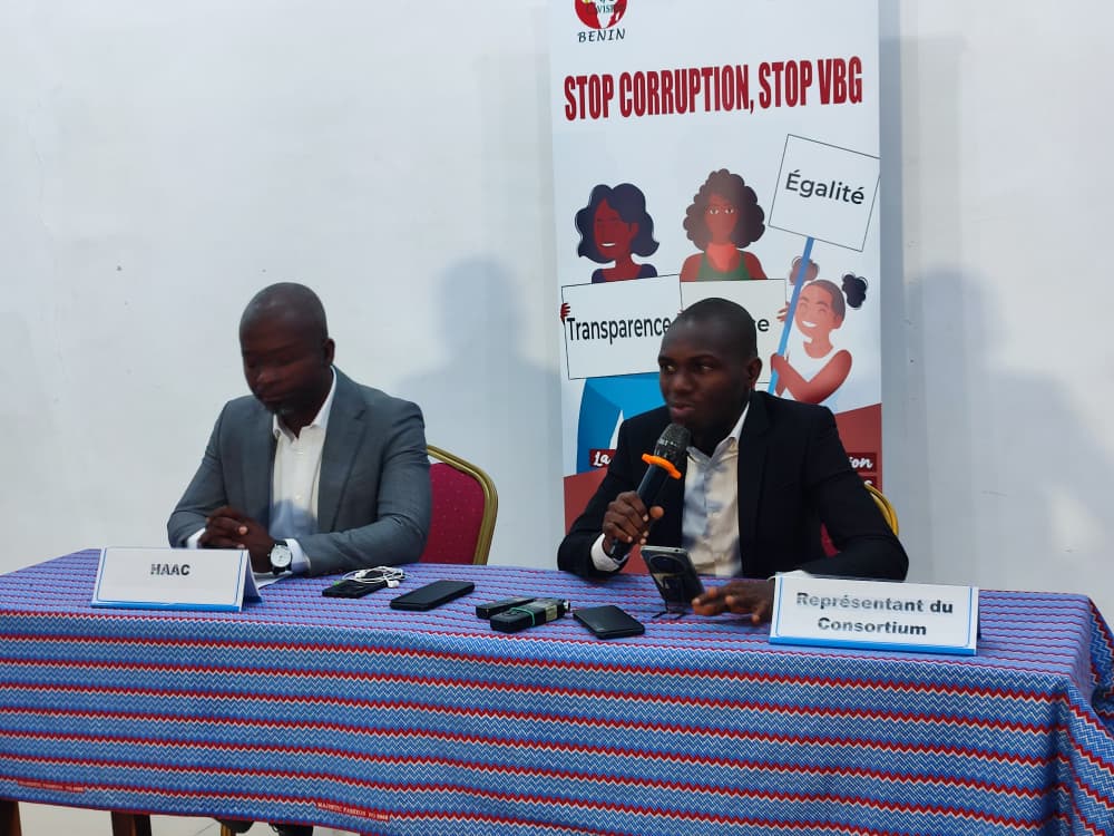 Deux hommes assis à une table avec micros et panneaux derrière eux sur le thème « Stop Corruption, Stop VBG