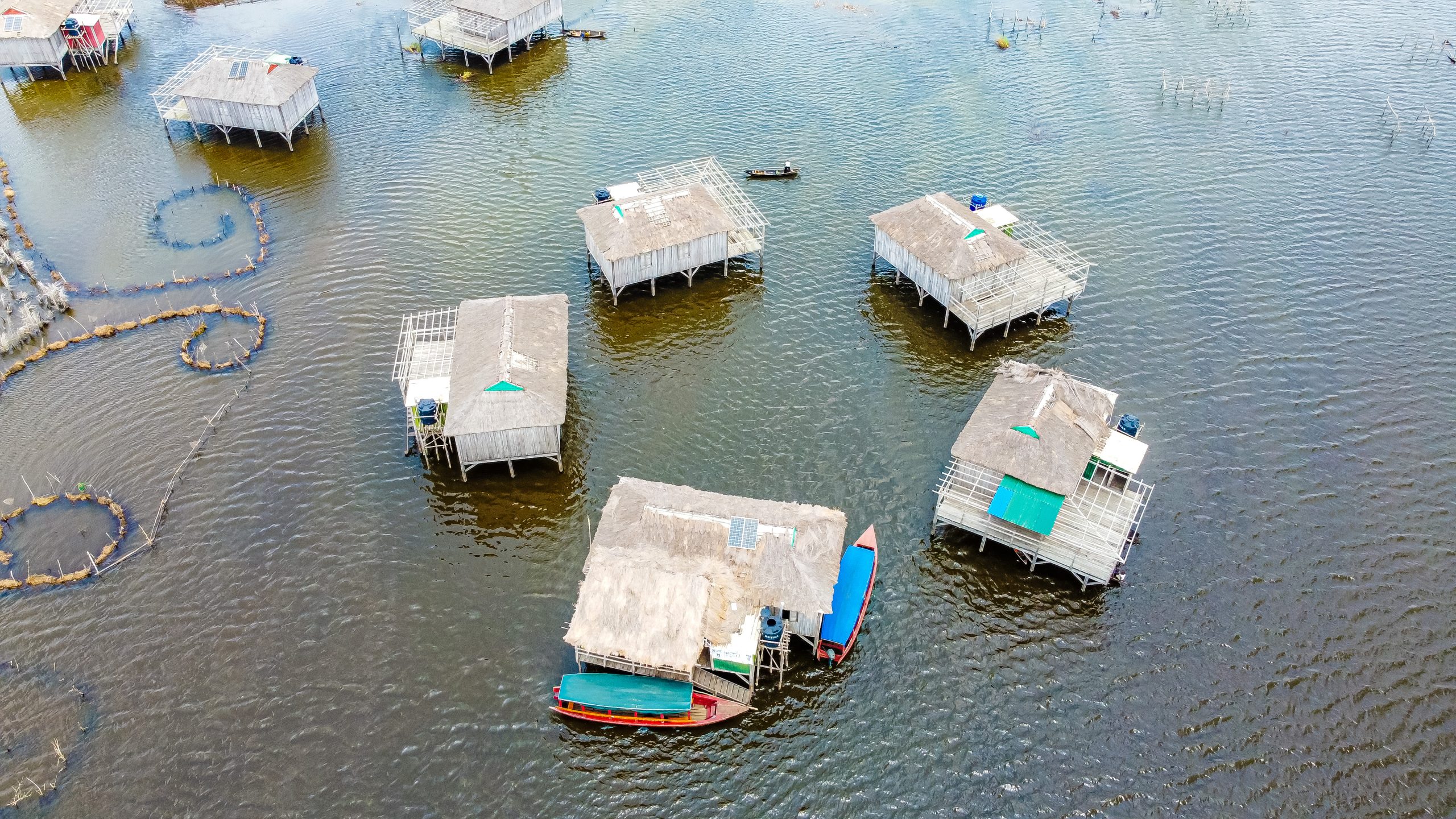 Maisons sur pilotis avec toits en chaume et petites embarcations sur une étendue d'eau calme vue aérienne