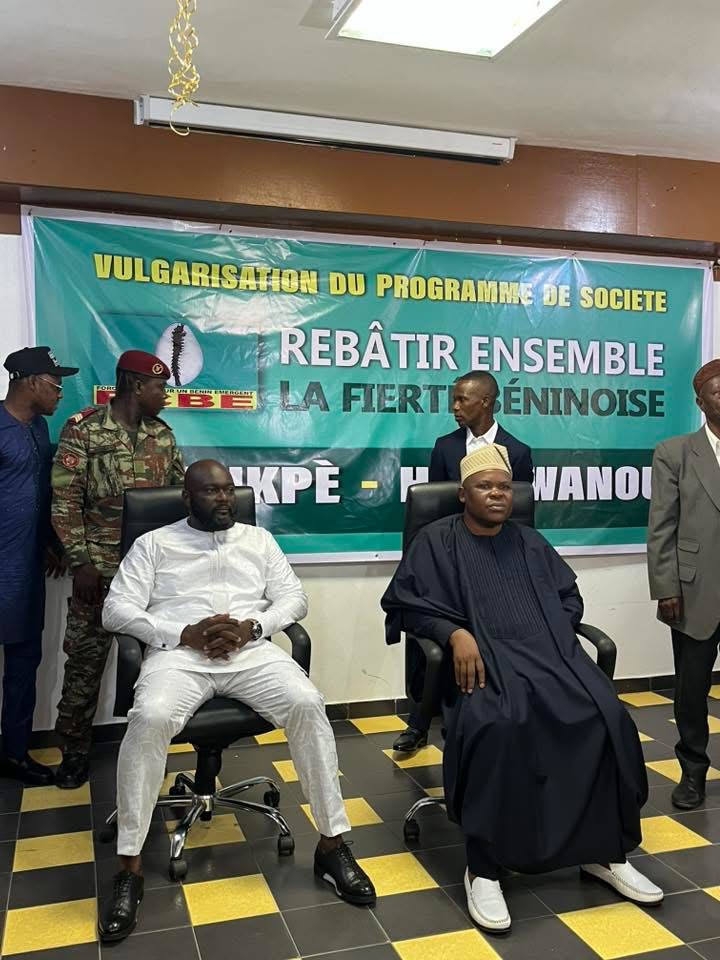 Deux hommes assis en costume traditionnel devant une banderole de programme de société entourés de plusieurs personnes debout