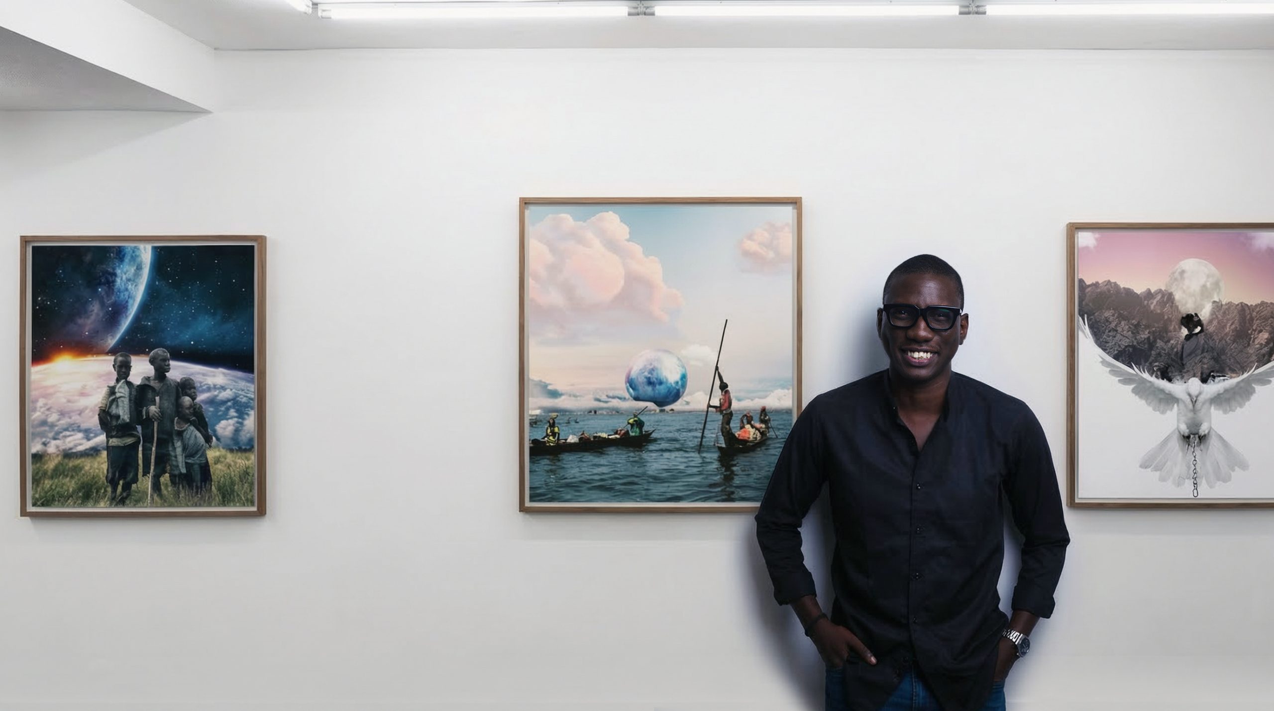 Un homme souriant en chemise noire pose devant trois tableaux accrochés dans une galerie d'art aux murs blancs