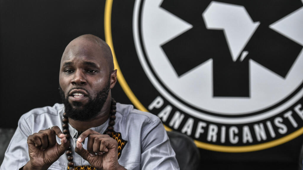Un homme barbu en chemise grise gesticule devant un logo rond noir et blanc avec un continent africain stylisé