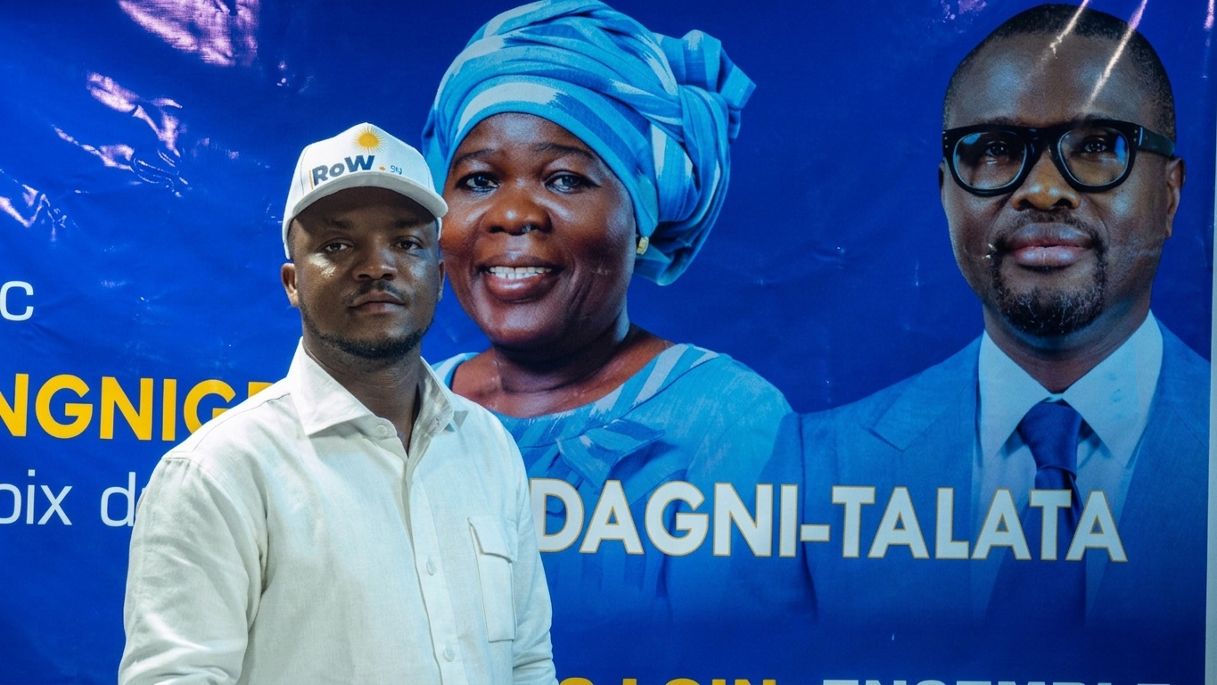 Homme en chemise claire et casquette blanche posé devant une grande affiche bleue avec deux portraits et du texte partiellement visible