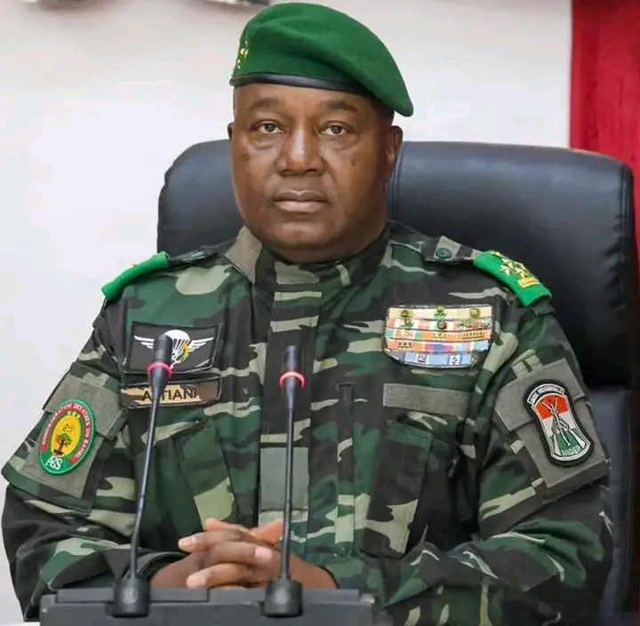 Un homme en uniforme militaire camouflage et béret vert est assis derrière un bureau avec deux microphones devant lui