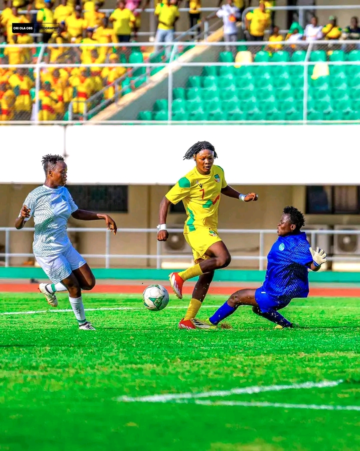 Une joueuse en maillot jaune dribble entre une adversaire et une gardienne en maillot bleu lors d'un match de football féminin
