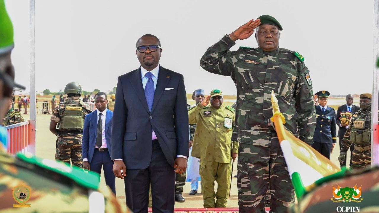 Un homme en costume sombre et un militaire saluent debout devant des soldats en uniformes face à eux