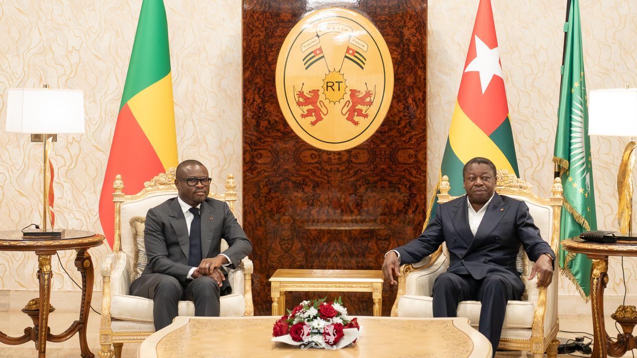 Deux hommes en costume assis dans un salon luxueux avec les drapeaux du Bénin, du Togo et de l'Union africaine en arrière-plan