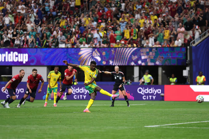 Un joueur en maillot jaune tire un penalty devant un gardien en maillot rouge sous les yeux des joueurs et de l'arbitre sur un terrain de football.