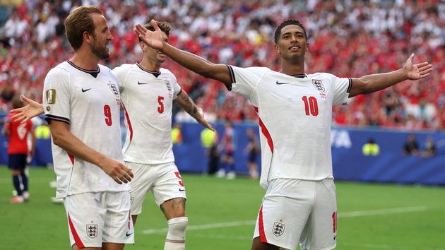 Trois joueurs en maillot blanc de football célèbrent un but sur un terrain avec foule en arrière-plan
