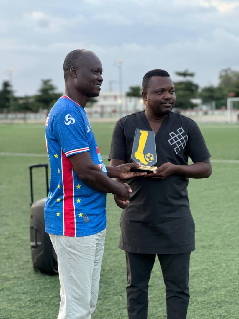 Deux hommes échangent une poignée de main, l’un tenant un trophée sur un terrain de football en plein air